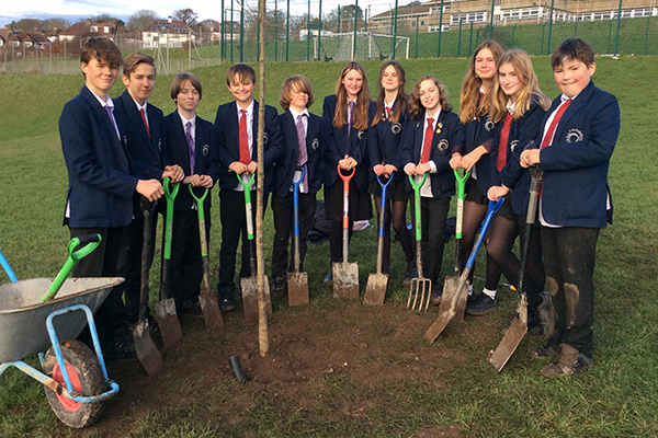 A group of school pupils are stood holding spades around a tree that they have just planted.
