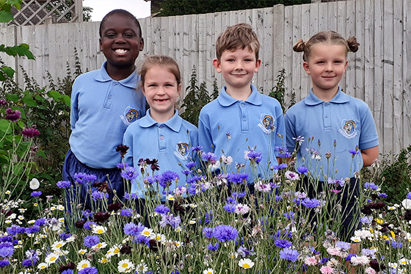 Young school students are stood in their school uniforms in front of a bed of flowers that they have planted.