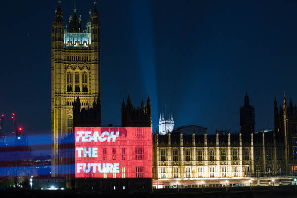 The houses of parliament lit up at night with a projection of 'Teach the future' written in white on a red background.